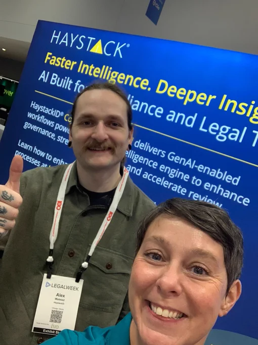 Two conference attendees posing with thumbs up in front of a Haystack AI technology booth at Legalweek event.