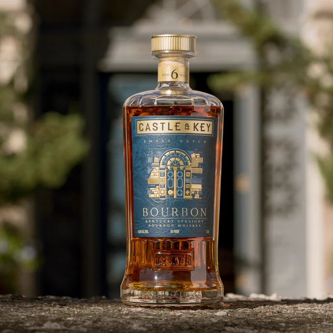 A bottle of Castle & Key Kentucky Straight Bourbon Whiskey is placed on a stone surface. The bottle features a blue and gold label with ornate design elements. A building's blurred exterior serves as the background.