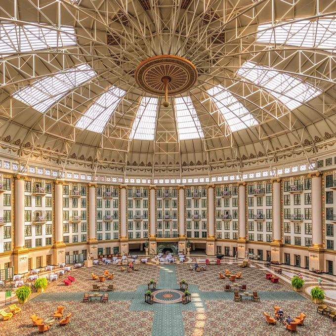 A large, elegant domed atrium of the West Baden Springs Hotel, featuring intricate details and a spacious open area with seating arrangements. The dome has a glass and metal framework allowing natural light to pour in. The floor is adorned with elaborate patterns and surrounded by green plants.