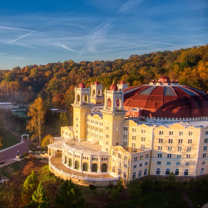 A grand, multi-story West Baden Springs Hotel with a large domed roof and two towers is set against a backdrop of lush, forested hills in autumn colors. The architecture is ornate, featuring numerous windows and a prominent entrance with columns. A road leads up to the building, and the sky is clear with wispy clouds.