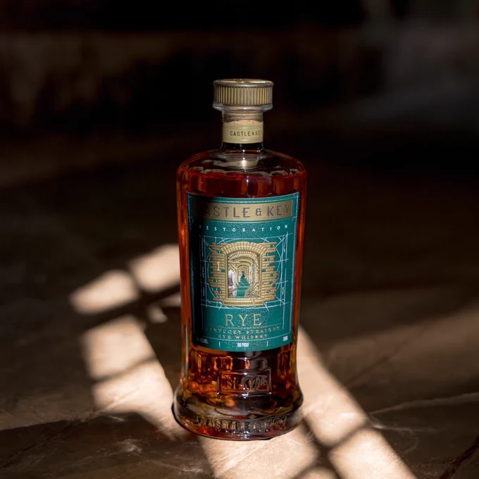 A bottle of Castle & Key Restoration Rye is placed on a wooden surface, with sunlight casting shadows across the scene. The bottle's label is visible, featuring intricate designs and a teal color scheme.