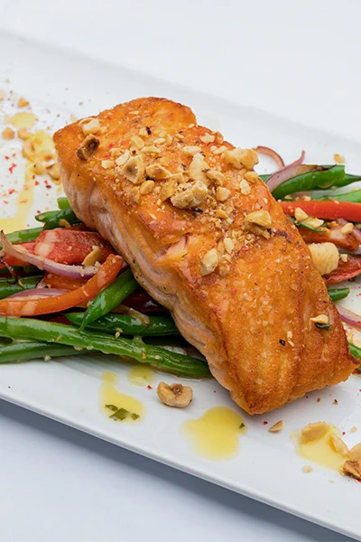 Piece of cooked salmon topped with hazelnuts on top of a green bean salad with a vinaigrette across the white plate 