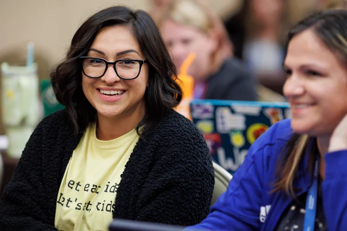 A photo of a woman smiling at the camera. She is wearing glasses and a black sweater. She is sitting with other people. 