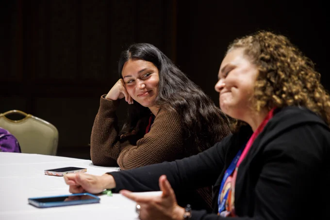 Photo of two women sitting at a table for discussion. One woman is reacting to the discussion, while the other woman is looking at the other and smiling. 