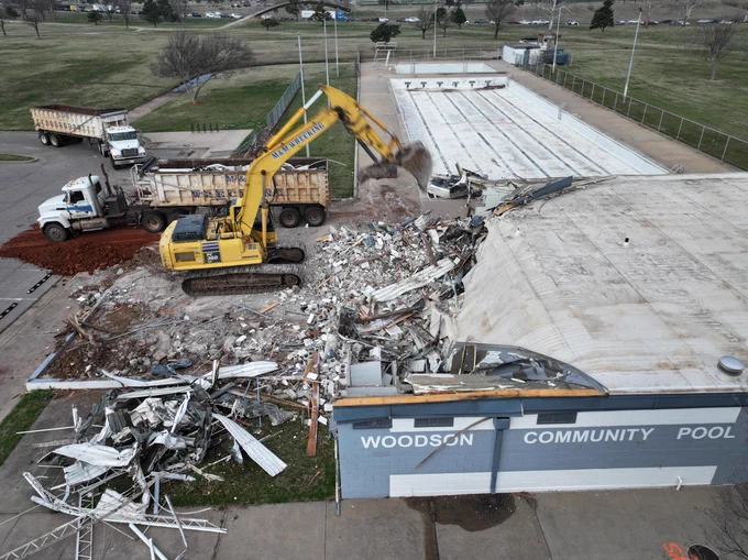 Yellow excavator demolishing the old structure beside an empty community pool with debris scattered around and dump trucks waiting to haul it away.