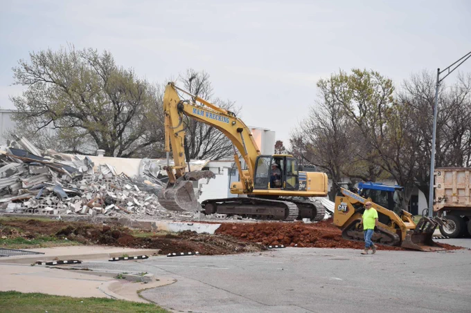 Yellow excavator clearing debris from a demolished building site with a worker nearby on a cloudy day.