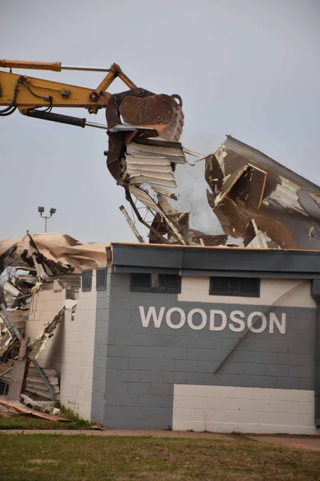 Excavator tearing down the roof and walls of a building labeled WOODSON during demolition.