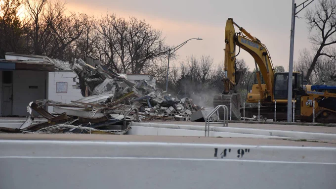 Yellow excavator demolishing a building near an empty outdoor swimming pool at sunrise.