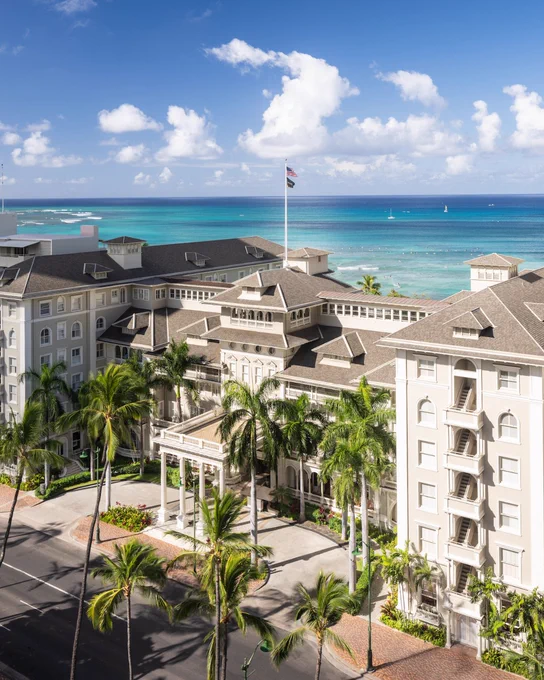 Aerial view of the Moana Surfrider hotel in Waikiki, with the ocean in the background. The historic building features a large entrance flanked by tall palm trees, under a clear blue sky.