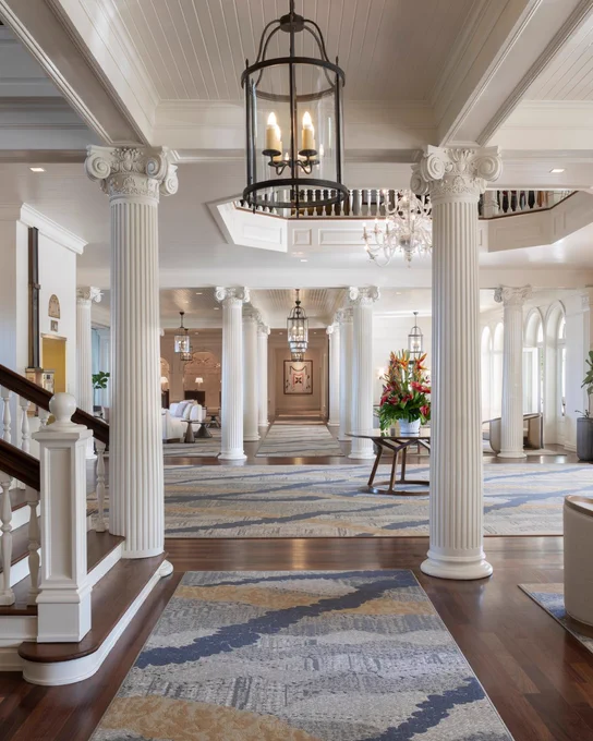 Luxurious hotel lobby with grand white columns and high ceilings, featuring elegant chandeliers and contemporary lighting fixtures. The space is adorned with abstract-patterned rugs on polished wooden floors, and vibrant flower arrangements add a touch of color. Stairs lead to an upper level, enhancing the opulent atmosphere.