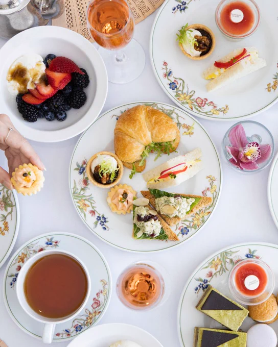 An elegant table setting with various foods on floral china plates. Dishes include a croissant sandwich, pastries, a bowl of berries with yogurt, and a cup of tea. Drinks in wine glasses and a small flower arrangement are present.
