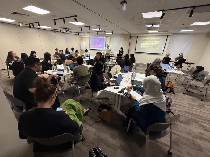 Room with people seated around tables, facing two projector screens.