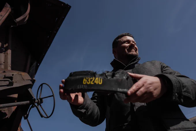 Serhii Kerekesha stands beside a freight wagon and holds a blackened, damaged metal fragment marked with yellow numbers. He raises the piece with both hands, showing it to the camera. Sunlight falls across his face as he looks slightly upward and smiles. Behind him, parts of a rusted railway wagon and metal mechanisms are visible against a clear blue sky, highlighting the industrial railway setting.