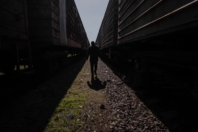 A railway worker walks between two long freight trains standing on parallel tracks. The person appears in silhouette against bright daylight, moving along the gravel path between the railcars as sunlight creates a narrow strip of light on the ground.