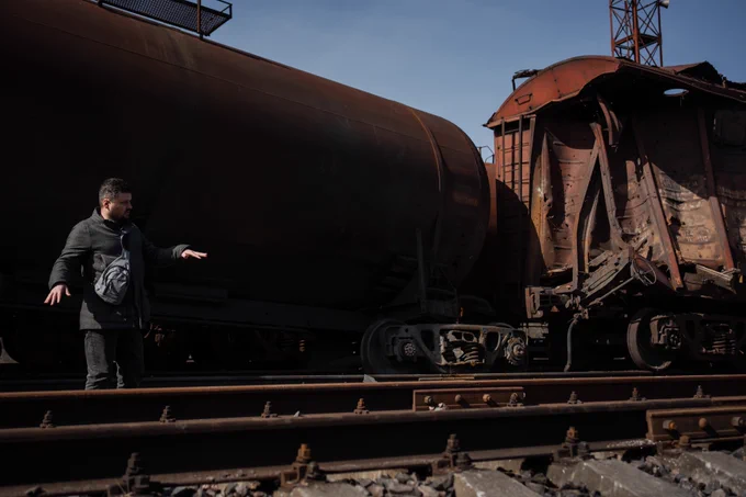 Serhii Kerekesha, Head of the Kakhovka Track Distance enterprise, stands on a railway line in Snihurivka, Mykolaiv Oblast. He wears a dark coat and a small crossbody bag while gesturing with his hand as if explaining something about the railway. Behind him stand two freight wagons: a large cylindrical tank wagon and another heavily damaged wagon with torn metal panels and visible holes. The rusty surfaces of the railcars contrast with the bright blue sky above the tracks.