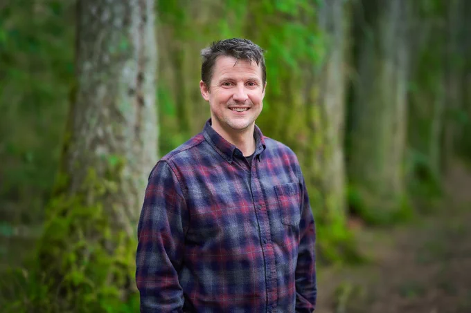 Man wearing a red and blue checked shirt standing in a dense forest with tall trees and green moss.