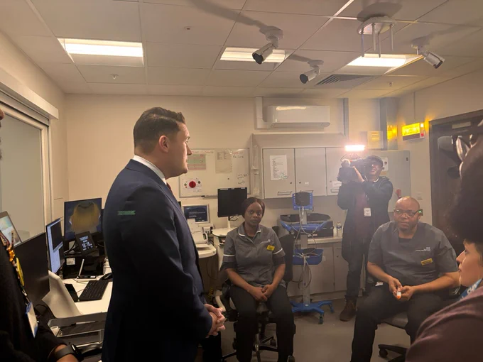 Wes Streeting in a suit speaking with hospital colleagues in the CT and MRI control room while a camera crew records the discussion.