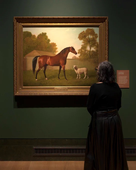 A person stands in front of a framed painting. The painting depicts a horse and a sheep in a pastoral setting.