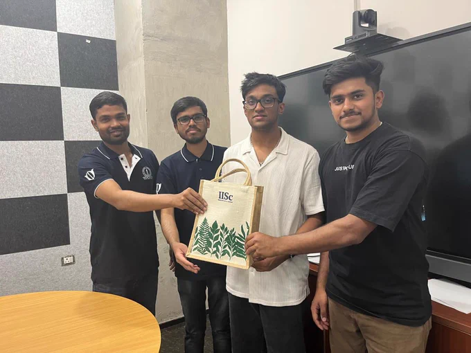Four young men standing indoors in a classroom or meeting room, jointly holding a tote bag with the IISc logo and plant illustrations. A round table is visible in the foreground, and a large screen with a camera mounted above it is behind them. One wall has a black-and-white checkered acoustic panel, and the group appears to be posing after receiving or presenting the bag.