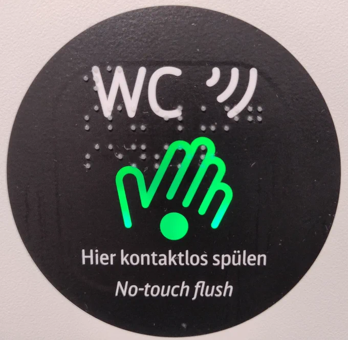 Sticker in einem WC eines ICE-Zugs: "Hier kontaktlos spülen". Mit eingraviertem Braille-Schriftzug.