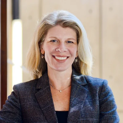 A smiling woman with shoulder-length blonde hair wearing a dark plaid blazer and small earrings, standing indoors with a softly lit beige background.