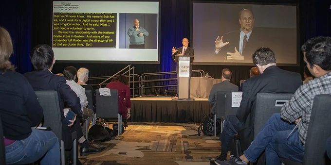 A man speaking on a stage behind a podium to participants at the C S U N Assistive Technology Conference.