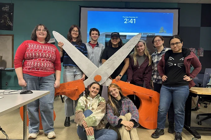 A group of nine students poses for a photo with a gigantic pair of scissors.