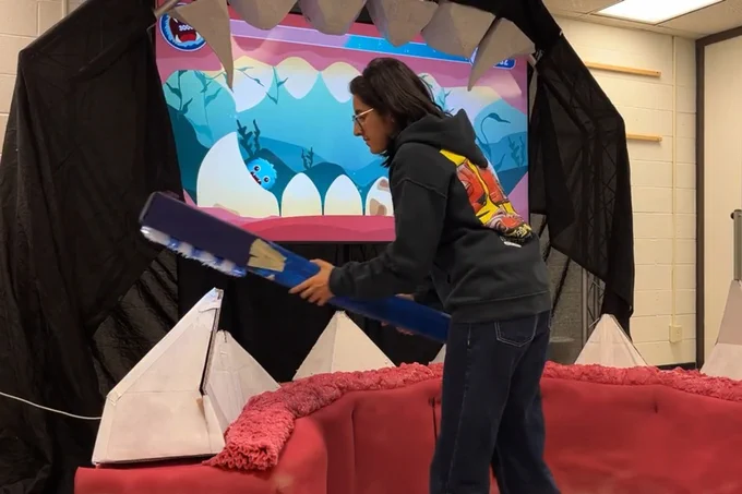 A student uses a gigantic toothbrush as a game controller on a large mouth and teeth. 