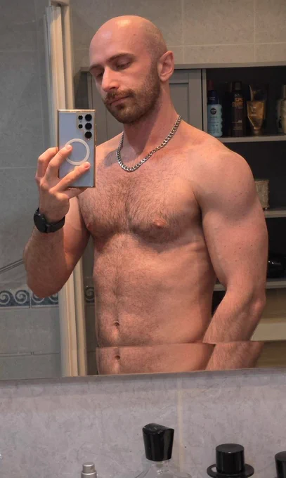 A bald man with a short brown beard and mustache takes a mirror selfie in a bathroom. He is shirtless, wearing a silver chain necklace and a black smartwatch, showing his hairy chest, toned shoulders, arms and abs. He holds a light blue smartphone in his right hand and looks down at the screen.
