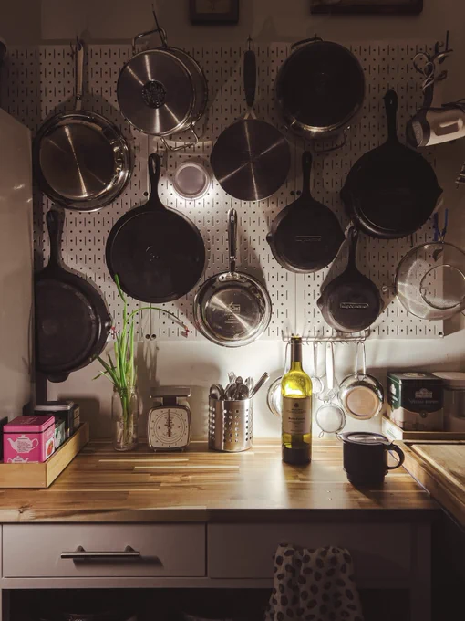 Wall Control metal pegboard mounted on a kitchen wall holding an organized display of cast iron and stainless steel pans, utensils, and kitchen tools above a wooden countertop with a scale, olive oil bottle, mug, and cooking accessories. Photo by customer Ania.