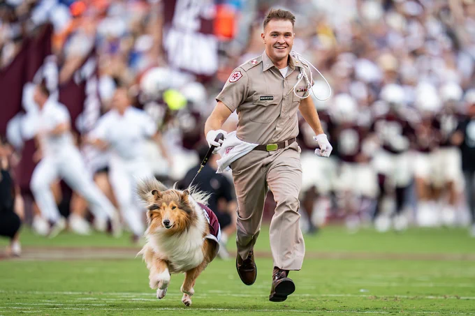 A person in a uniform runs joyfully with Reveille, the Texas A&M University mascot, on a football field. The background shows a lively crowd.
