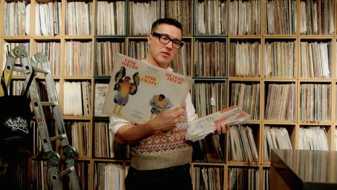 Jonny Trunk holding vinyl records in front of a wall of LP shelves.