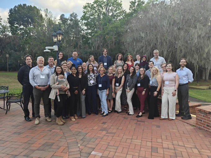 Large group photo of current and alumni NMPT and BREATHE T32 trainees standing together outdoors on a brick patio. The group is arranged in several rows and wearing conference name badges, smiling at the camera. Trees with Spanish moss and a lamppost are visible in the background, and one person in the front row is holding a baby.
