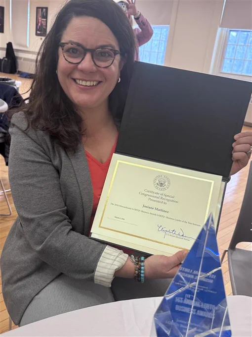 Josiane Martinez, a Latina woman with shoulder-length dark hair wearing a grey blazer and tortoise-shell glasses, smiles while she displays her certificate for an award.