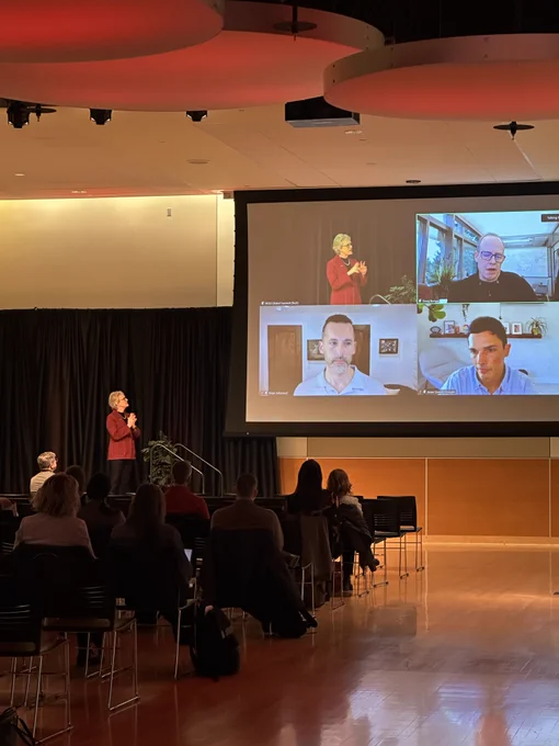 President Cantwell stands on a stage addressing an audience, with a large screen behind her showing multiple remote participants during the Global Summit. 
