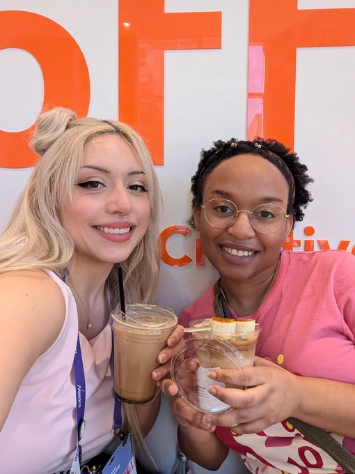 Us smiling, wearing pink shirts and holding the drinks. I'm on the left with long blonde hair and hair buns. Claire is on the right with fluffy dark brown hair and golden ring hair jewelry.