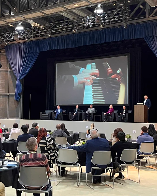 Abe Dyck on stage presenting to an audience. On the screen is an image of someone playing a piano.