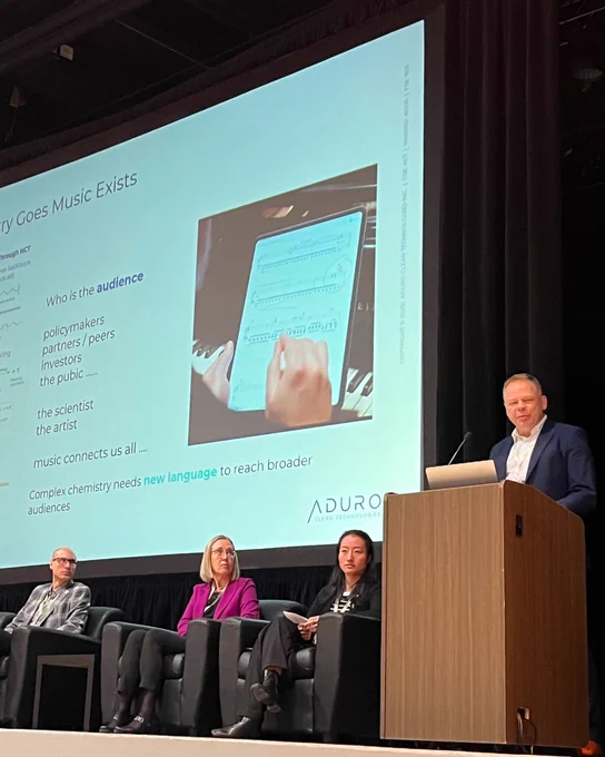 Abe Dyck on stage behind a podium presenting with a presentation screen in the background and other panelists on stage.
