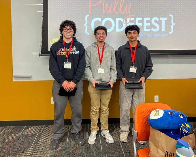 Three young participants wearing event badges stand in front of a Philly Codefest presentation screen holding black boxes as awards.