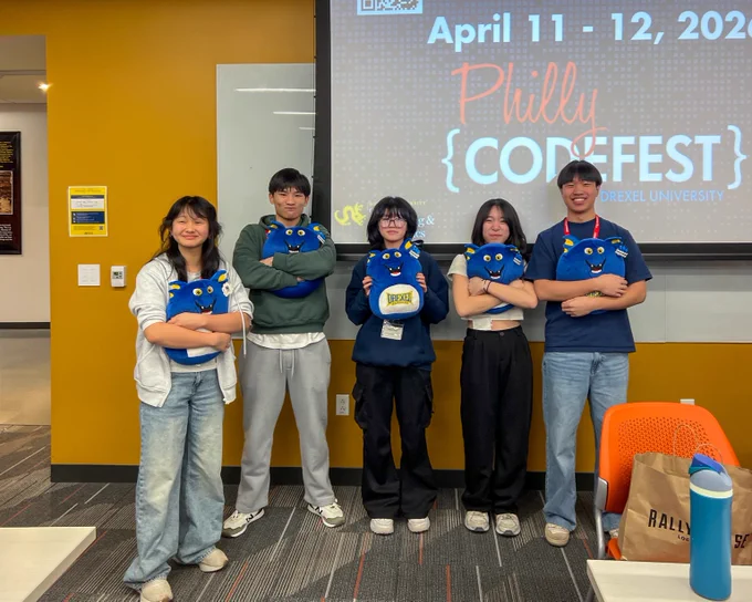 Five participants stand in a row holding blue plush toys shaped like monsters at Philly CodeFest event in front of a presentation screen.