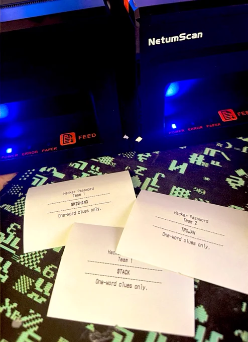 Two black NetumScan thermal receipt printers with blue power indicator lights, sitting on a pixelated hacker-themed mat. In front of them, three freshly printed receipts display word cards for a game called “Hacker Password” — Team 1 has the words SMISHING and STACK, Team 2 has TROJAN — each with the instruction “One-word clues only.” The setup suggests a live game show in progress, with printed clue cards being dispensed in real time.