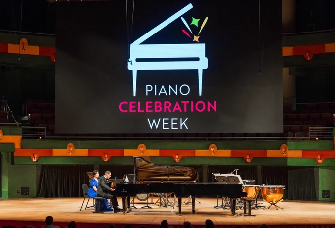 A person is playing the piano on stage with another person turning pages. Behind them, a large screen displays the words "Piano Celebration Week" with a stylized piano graphic. The stage is set with percussion instruments in a concert hall at TAMU-CC.