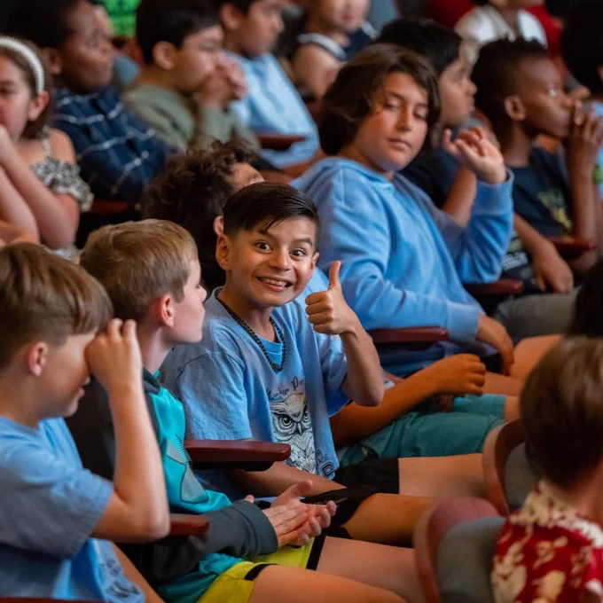 A group of children seated in an auditorium on the campus of Texas A&M University-Corpus Christi. One child in the center is smiling and giving a thumbs-up gesture. The rest are focused on a performance or speaker.