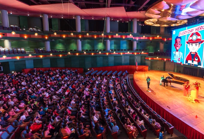 A large auditorium at the Performing Arts Center on the campus of Texas A&M University-Corpus Christi filled with people seated in rows. On stage, a person stands next to a grand piano, addressing the audience. A large screen displays animated characters under the headline "Everyday Heroes." The theater features multiple tiers of seating and warm lighting.