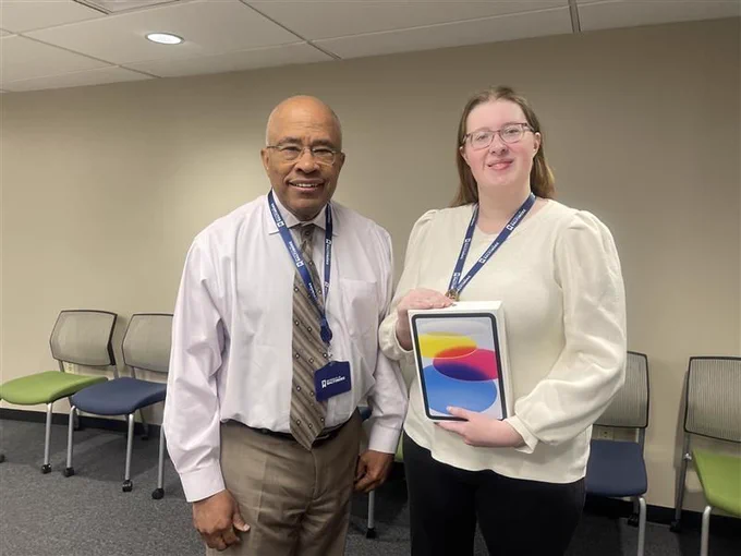 One student and President Kurt Schmoke  pose together indoors while a ipad the student received as a prize.