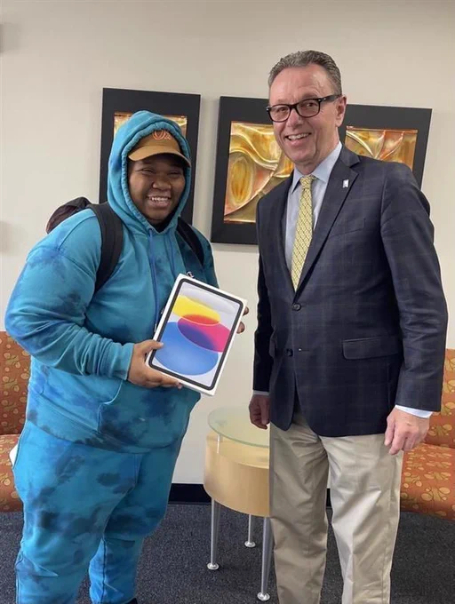 A smiling student in a blue hoodie holds a new iPad while standing next to a university administrator in a suit inside an office space.