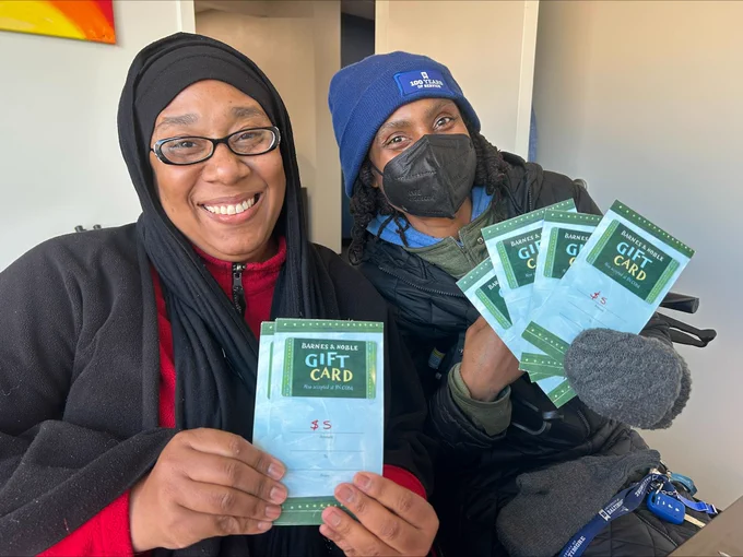 Two smiling individuals sit together holding Barnes & Noble gift cards, celebrating receiving the cards indoors.