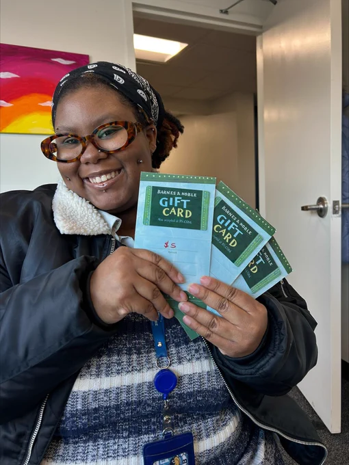 A smiling person wearing glasses holds several Barnes & Noble gift cards while sitting indoors.
