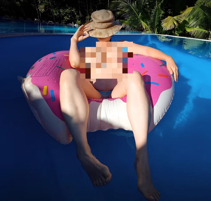 The image shows a person sitting on a large, pink donut-shaped pool float in a swimming pool. The individual is wearing a straw hat that covers their face and has a pixelated blur effect over their torso. They are positioned in the center of the pool, surrounded by blue water, with palm trees visible in the background under bright, sunny lighting.