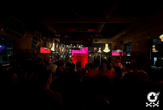 Photo of room at Brazen Head pub in Toronto for TFC 2026 Supporters Forum. Supporters standing look on towards glowing pink/red stage backdrop with player/coach guests speaking.. Red Patch 20-year watermark logo in bottom corner.